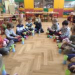 Kilkanaścioro dzieci siedzi w siadzie skrzyżnym na podłodze w sali lekcyjnej. Dzieci trzymają w rękach kolorowe kubeczki plastikowe. Przed każdym dzieckiem stoi wieża zbudowana z dwóch kubeczków