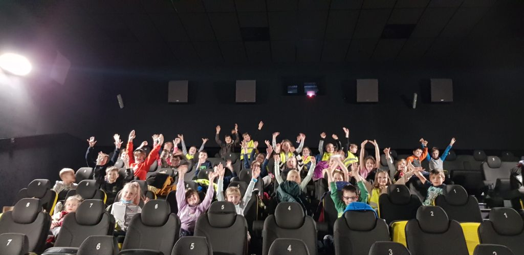 Grupa około 35 dzieci siedzi na fotelach w sali kinowej. Dzieci z uśmiechem na twarzy trzymają ręce w górze.