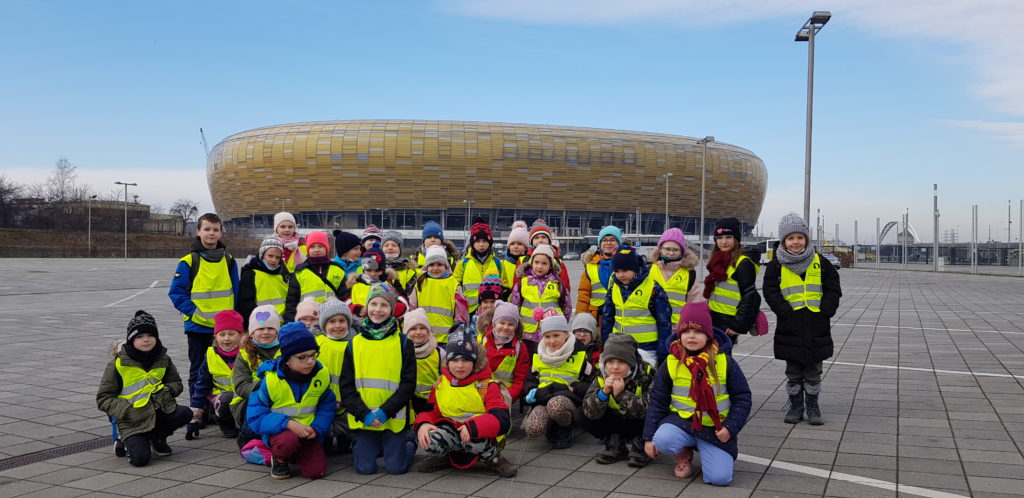 Trzydziestu pięciu uczniów ubranych w żółte kamizelki ustawionych w trzech rzędach na tle stadionu Polsat Plus Arena Gdańsk