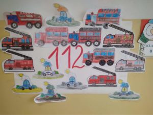 Na ścianie wisi plakat z numerem alarmowym 112 oraz z pojazdami służb ratunkowych. 