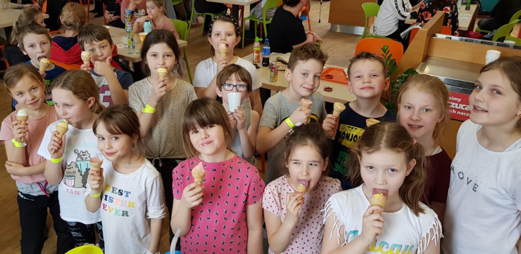 Grupa ok. 15 dzieci z uśmiechem na twarzy ze smakiem je lody.