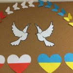 Dwa białe gołąbki otoczone od góry motylami ułożonymi w półkole w kolorach flag Polski i Ukrainy. Na dole serca w kolorach flag: biało-czerwone oraz niebiesko-żółte
