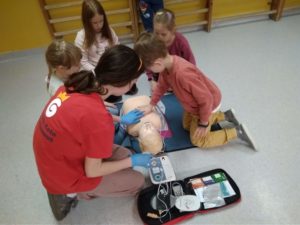 Uczennica Koła HOPR pomaga pierwszoklasiście wykonać uciski klatki piersiowej na fantomie. Na pierwszym planie podłączony do fantoma defibrylator treningowy AED. W tle widać 4 przyglądających się uczniów.