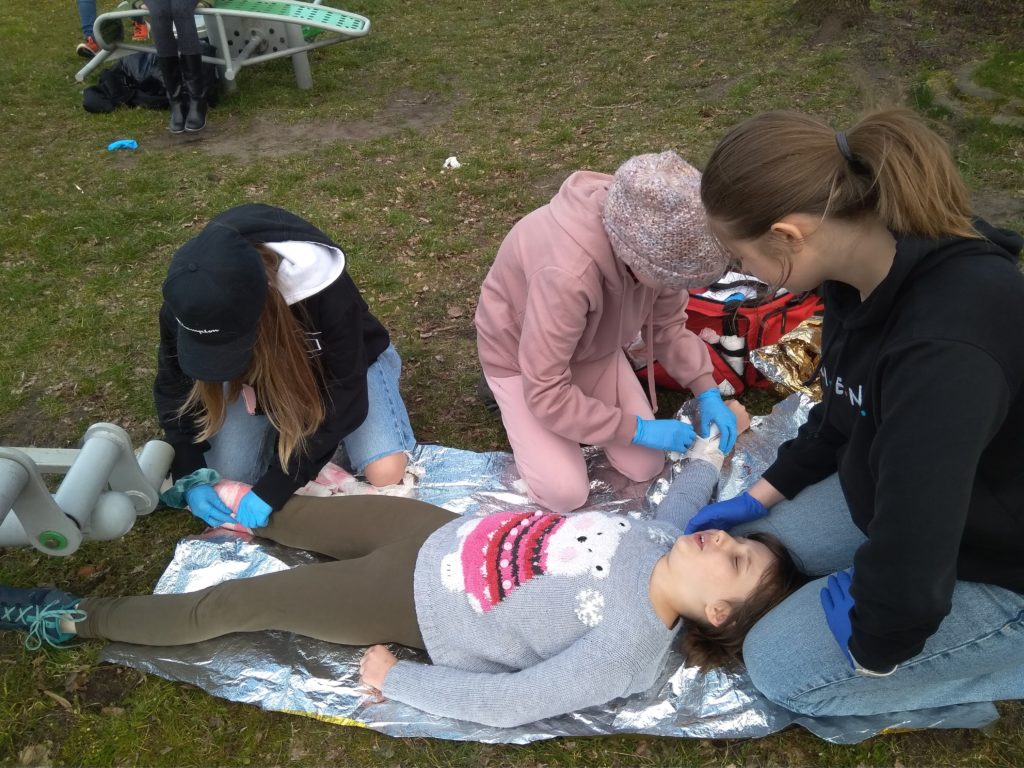 Jedna z uczennic symuluje ranną leżąc na kocu termicznym na terenie szkolnego podwórka. 3 starsze uczennice udzielają jej pierwszej pomocy opatrując rany.