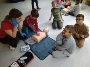 Pierwszoklasista ćwiczy masaż serca na fantomie z podłączonymi elektrodami AED. Uczennica koła HOPR symuluje wzywanie pomocy z innym uczniem. W tle przyglądający się uczniowie.