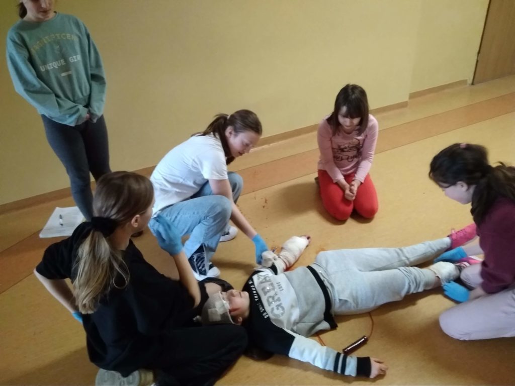 Na szkolnym korytarzu leży uczennica pozorująca krwotok. 3 uczennice starszych klas tamują krwotok i opatrują rany, dwie uczennice przyglądają się sytuacji.