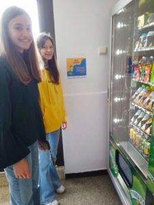 Przed szkolnym automatem z przekąskami stoją dwie uczennice 8 klasy i podejmują decyzję o zakupie napoju. W tle na ścianie wisi plakat z hasłem: Pij wodę!