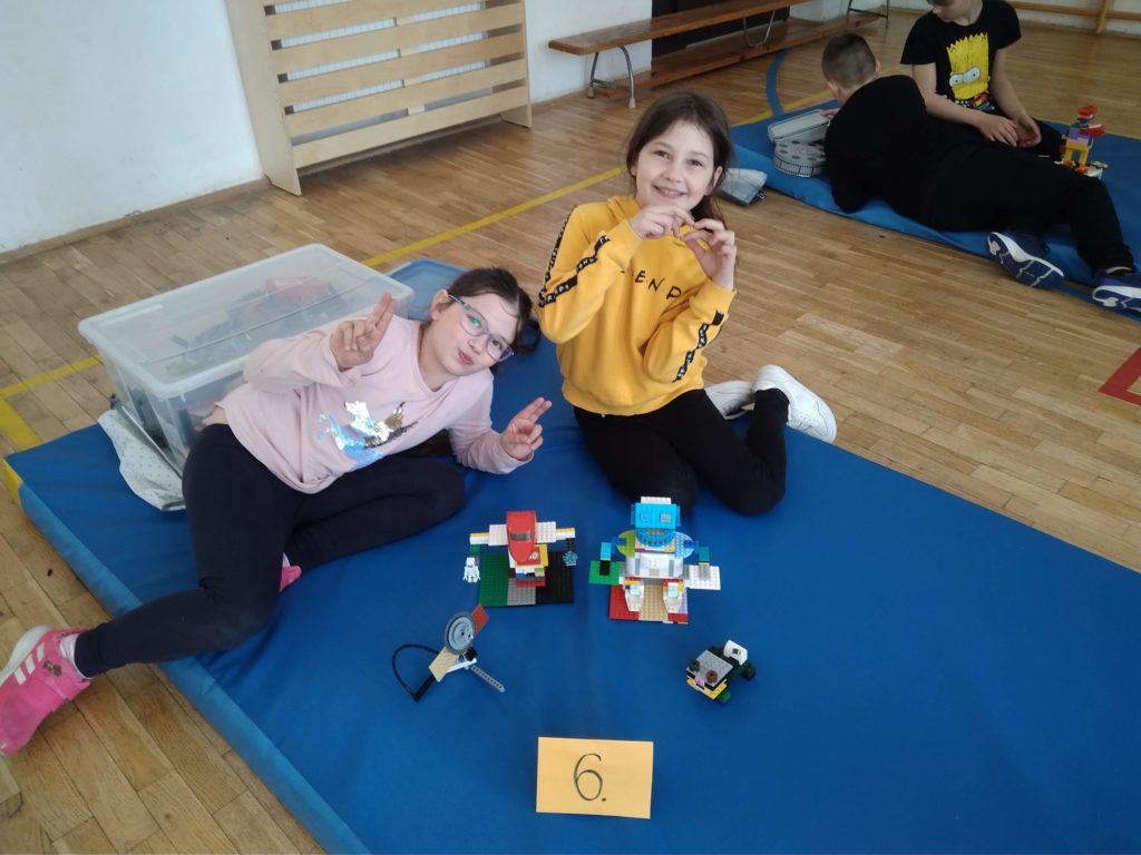 Na niebieskim materacu siedzą dwie uczennice, przed nimi leży ułożona przez nie budowla z klocków LEGO- roboty, w tle widać kolejny zespół uczniów i pudełka z klockami.