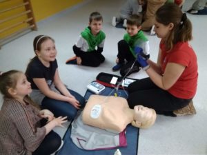 Uczennica Koła HOPR demonstruje czterem uczniom klasy pierwszej jak właściwie przykleić elektrody defibrylatora AED do klatki piersiowej fantoma.