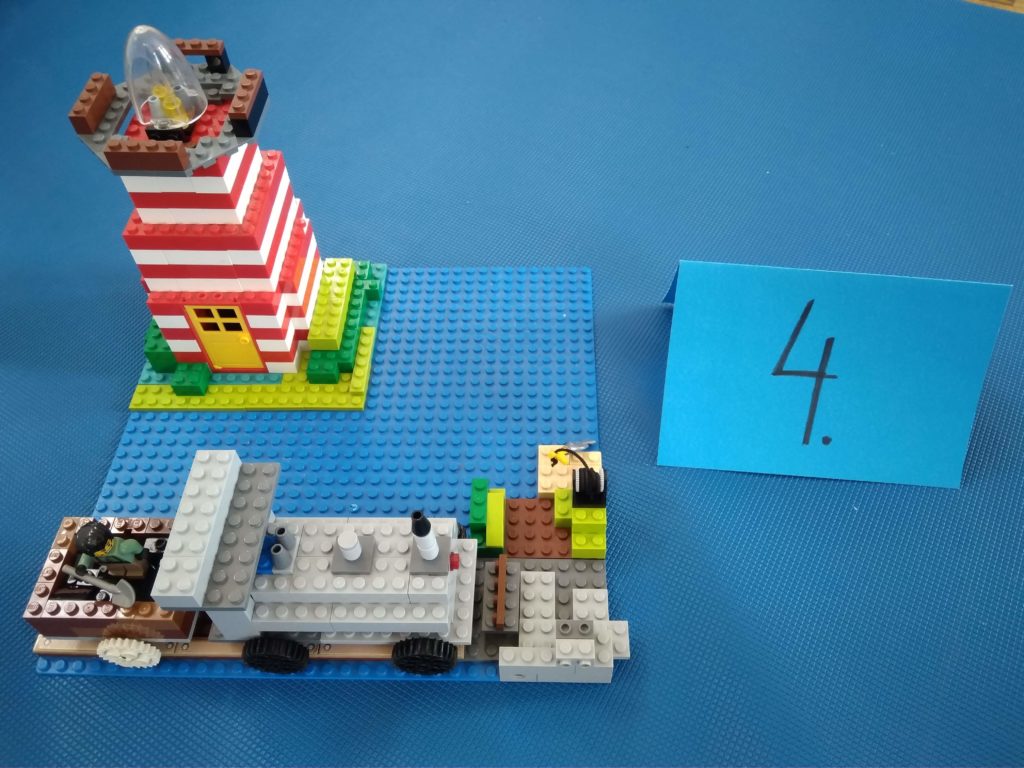 Na tle niebieskiego materaca stoi konstrukcja z klocków LEGO przedstawiająca nowoczesną latarnię morską i nowoczesny pojaz