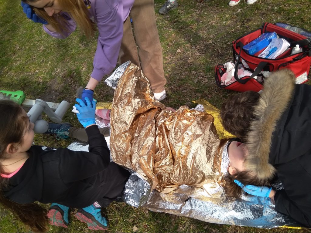 Pozorantce leżącej na trawie i okrytej kocem termicznym dwie uczennice opatrują rany, a jeden z uczniów sprawdza oddech. W tle apteczka pierwszej pomocy.