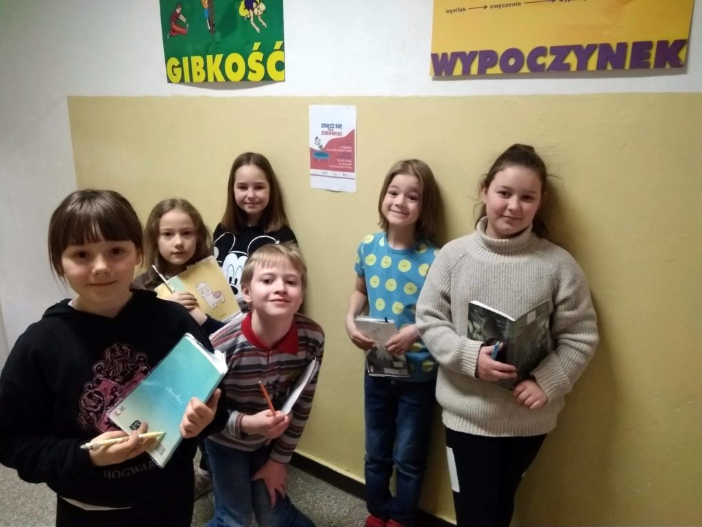 6 uczniów drugiej klasy trzyma w rękach notatniki i stoi przy wiszących plakatach promujących aktywność fizyczną.