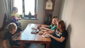 Cztery dziewczynki siedzą przy brązowym stole. Na stole leżą prostokątne przedmioty-foremki, łopatka oraz naczynie z patykami. W tle stoi zegar oraz jest okno