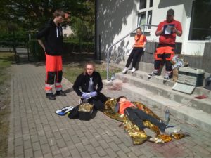 Na chodniku leży poszkodowany, urazowy chłopiec, któremu udziela pomocy uczennica z SP 9 w Wejherowie. Drużynie przyglądają się sędziujący ratownicy i pozorująca uczennica.