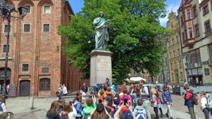 Uczniowie klasy IV b i c ustawieni tyłem w grupie, niektórzy z nich mają założone plecaki, w tle widać pomnik Mikołaja Kopernika oraz duże zielone drzewo, po bokach zarys zabytkowych budynków.