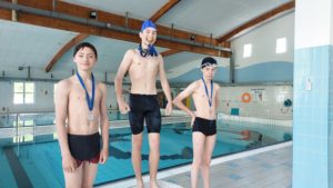 Na podium stoi trzech chłopców ubranych w czarne kąpielówki. Dwóch z nich ma medale zawieszone na szyi, a trzeci trzyma je w ustach. W tle są baseny pływackie.