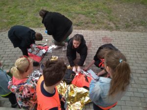 Na chodniku leży poszkodowany, urazowy chłopiec, któremu udziela pomocy trzyosobowa drużyna z SP 16 w Gdańsku. W tle otwarta apteczka i pozorujący uczniowie.