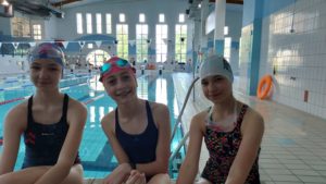 Trzy uśmiechnięte dziewczynki ubrane w stroje pływackie, z czepkami na głowie, siedzą w szeregu. Za nimi jest duży basen pływacki.