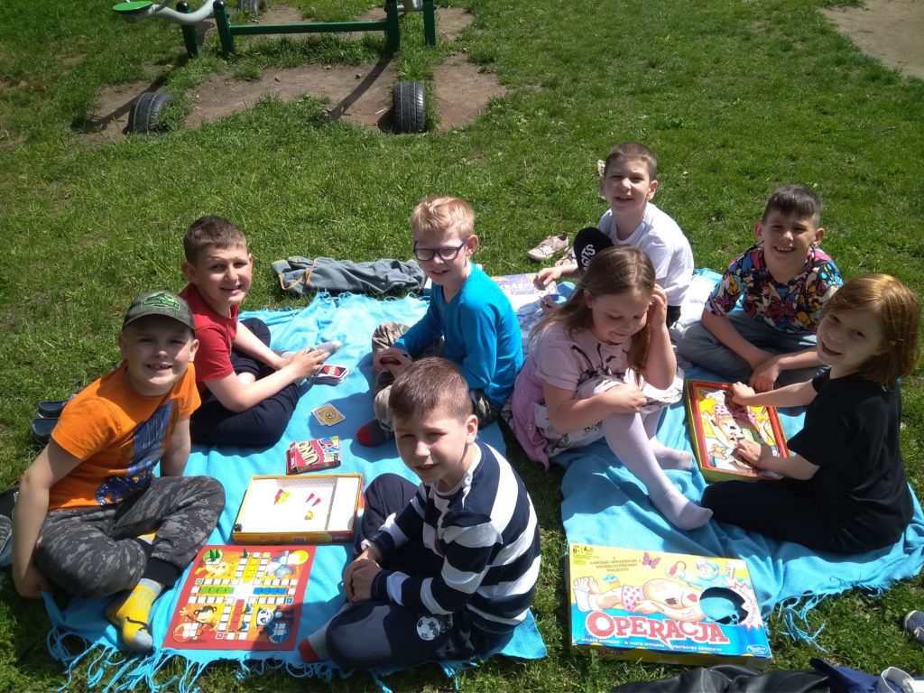 8 uczniów klasy 1c siedzi na kocykach w szkolnym ogródku i gra w gry planszowe.