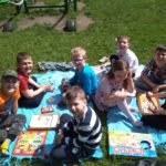 8 uczniów klasy 1c siedzi na kocykach w szkolnym ogródku i gra w gry planszowe. 