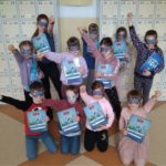 11 uczniów Koła Pierwszej Pomocy w maskach superbohaterów pozuje do zdjęcia trzymając w dłoni zeszyty ćwiczeń projektu FAST Heroes. W tle widać szkolne szafki na korytarzu. 
