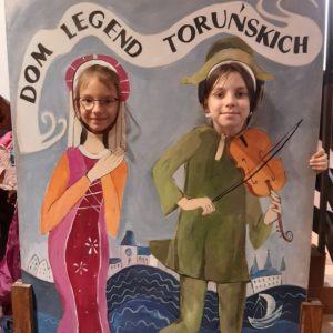  Dwie dziewczynki pozują do zdjęcia wystawiająć głowy z makiety domu legend toruńskich przedstawiajacej średniowieczną mieszczkę i grajka-skrzypka. W tle makiety Wisła i panorama Torunia. 