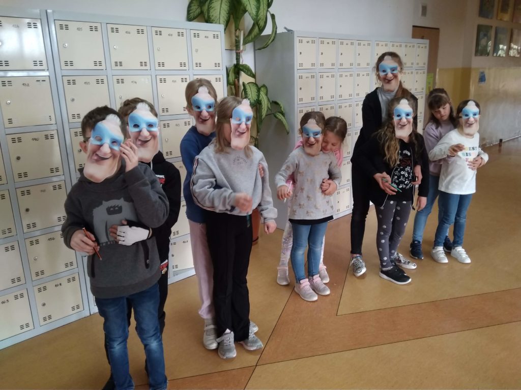 11 uczniów Koła Pierwszej Pomocy w maskach superbohaterów pozuje do zdjęcia trzymając w dłoni zeszyty ćwiczeń projektu FAST Heroes. W tle widać szkolne szafki na korytarzu. 