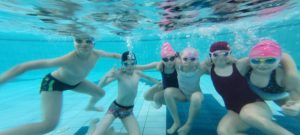 Sześcioro uczniów kl2a pod wodą pozuje do zdjęcia. Każdy z nich dmucha powietrze nosem ma założone okulary pływackie i czepek. 4 z prawej to dziewczynki w strojach jednoczęściowych a dwóch z lewej to chłopcy w kąpielówkach