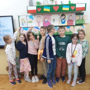 Uczniowie stoją pod tablicą, do której przyczepione magnesami są wykonane przez nich portrety - reprodukcje polskich malarzy - narysowane pastelami.
