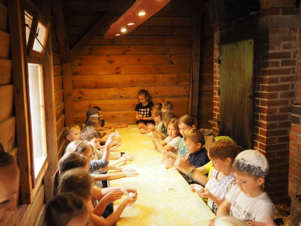Uczniowie siedzą po obu stronach bardzo długiego stołu, na którym leży żółta cerata. Na ceracie na środku stołu jest rozsypana mąka.