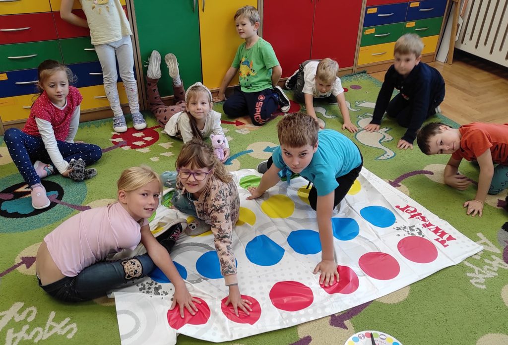 3 dzieci gra w grę Twister. W tle 6 dzieci siedzi na dywanie.