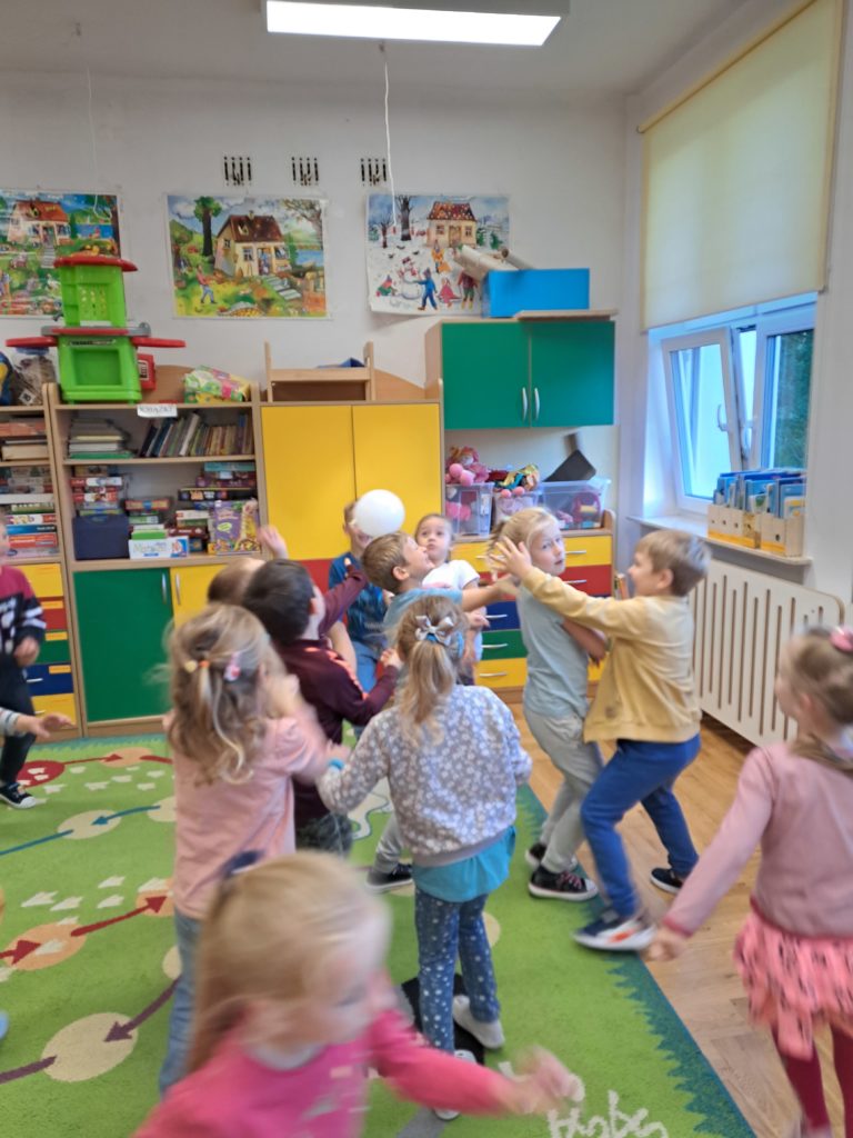 Dzieci poruszają się po sali podrzucając kremowy balon tak by nie spadł na podłogę. 