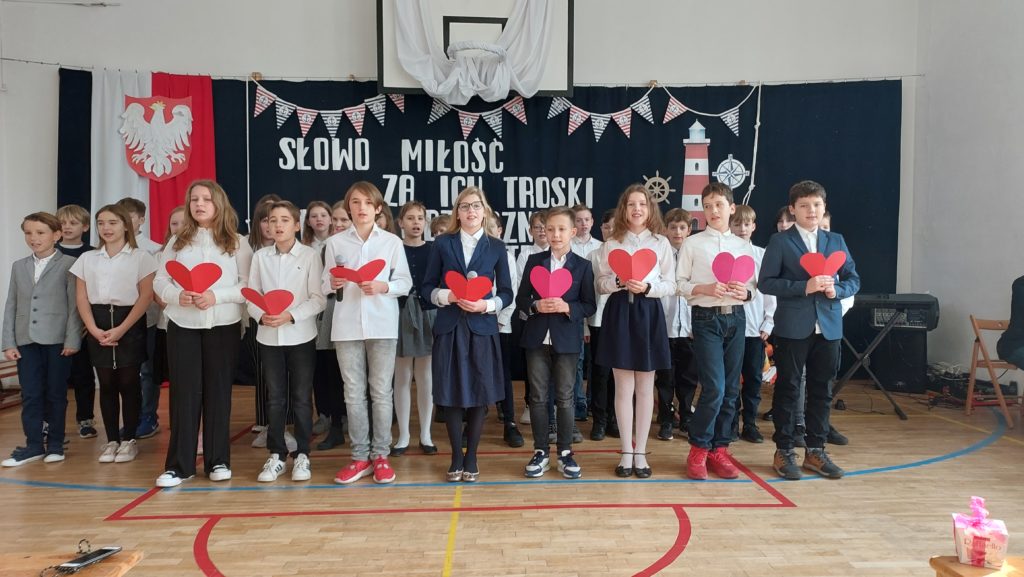 Uśmiechnięci uczniowie ubrani na galowo stoją w kilku szeregach. W pierwszym trzymają w rękach czerwone serduszka. W tle jest dekoracja: flaga i godło Polski, napis.