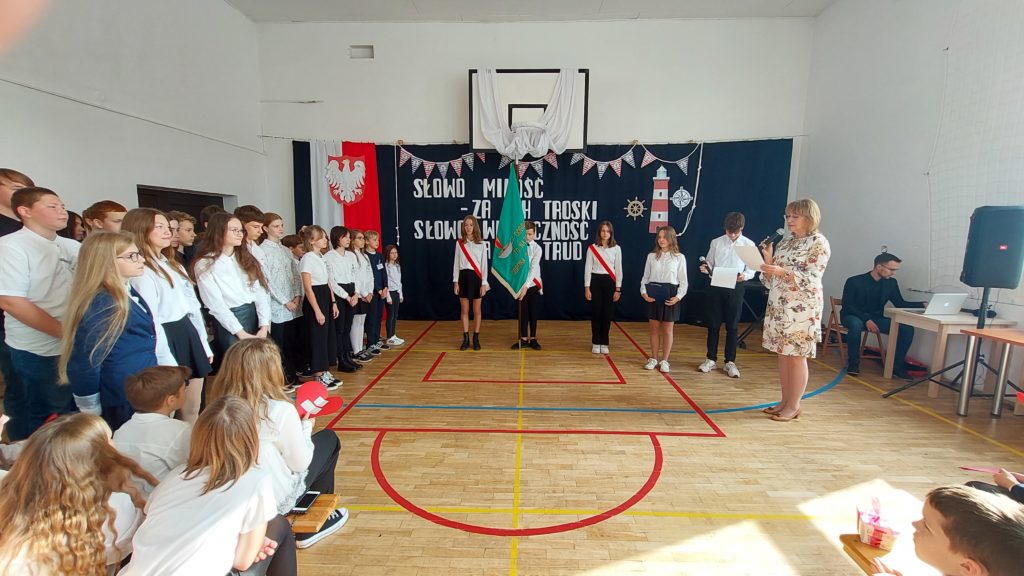 Pięcioro uczniów stoi w szeregu. Jeden z nich trzyma w rękach sztandar szkoły w kolorze zielonym. Po prawej stronie stoi pani dyrektor szkoły, a po lewej uczniowie ubrani na galowo. W tle jest dekoracja: flaga i godło Polski, napis.