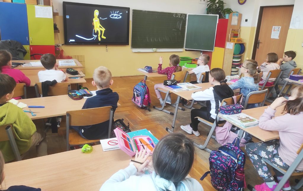 W sali lekcyjnej siedzą uczniowie klasy 2 obróceni przodem do tablicy multimedialnej, na której wyświetlony jest kadr filmu o udarze mózgu. 