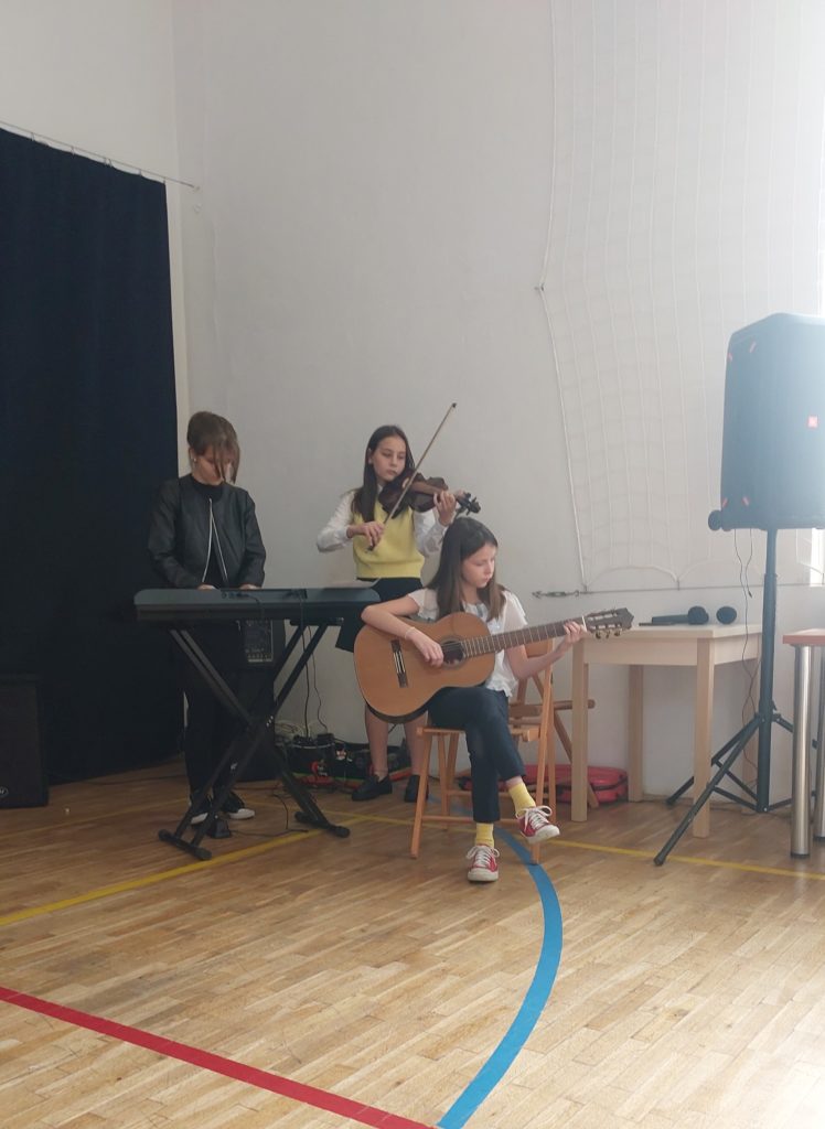 Dziewczynka ubrana na czarno stoi za keyboardem, druga dziewczynka stoi obok niej i trzyma w rękach oparte o szyję skrzypce. Trzecia dziewczynka siedzi na krześle i trzyma w rękach gitarę.