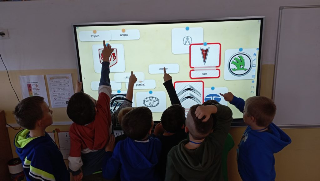 8 chłopców z klasy 1c stoi w sali lekcyjnej zwróconych twarzą do tablicy multimedialnej, z czego 4 z nich wskazuje palcem obrazki z wyświetlanego zadania.