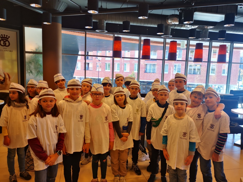 Uczniowie stoją w gromadzie i prezentują stroje. Dzieci ubrane są w białe koszulki z logo cukierni Pellowski. Na głowie mają czapeczki ochronne.