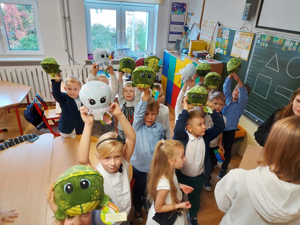 Grupa dwunastu uczniów z klasy "0" stoi blisko siebie na środku sali trzymając w rękach uniesionych nad głową maskotki w kształcie żółwia i meduzy. Na ich twarzach jest uśmiech.