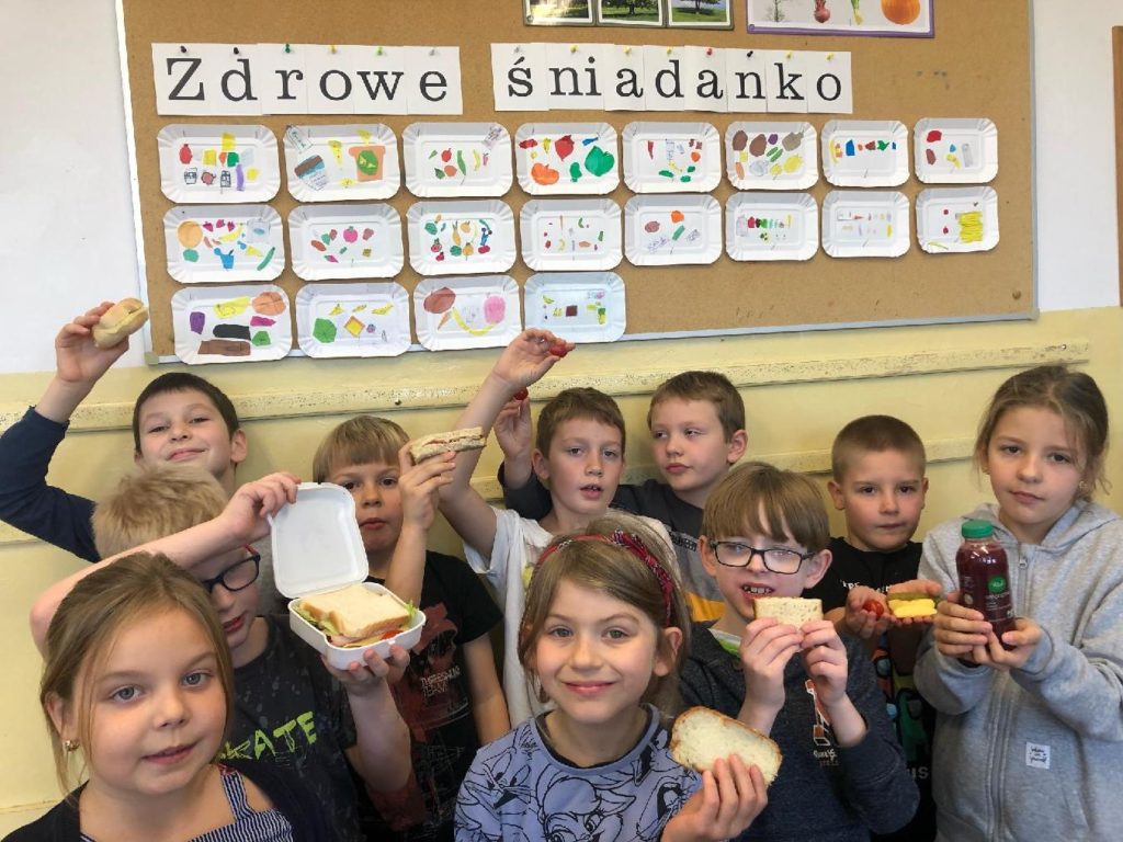 10 dzieci stoi przed tablicą. W rękach trzymają chleb, warzywa, owoce i soki. Ponad głowami uczniów widać napis "Zdrowe śniadanko" i prace plastyczne przedstawiające propozycje śniadania wyklejone z papieru.