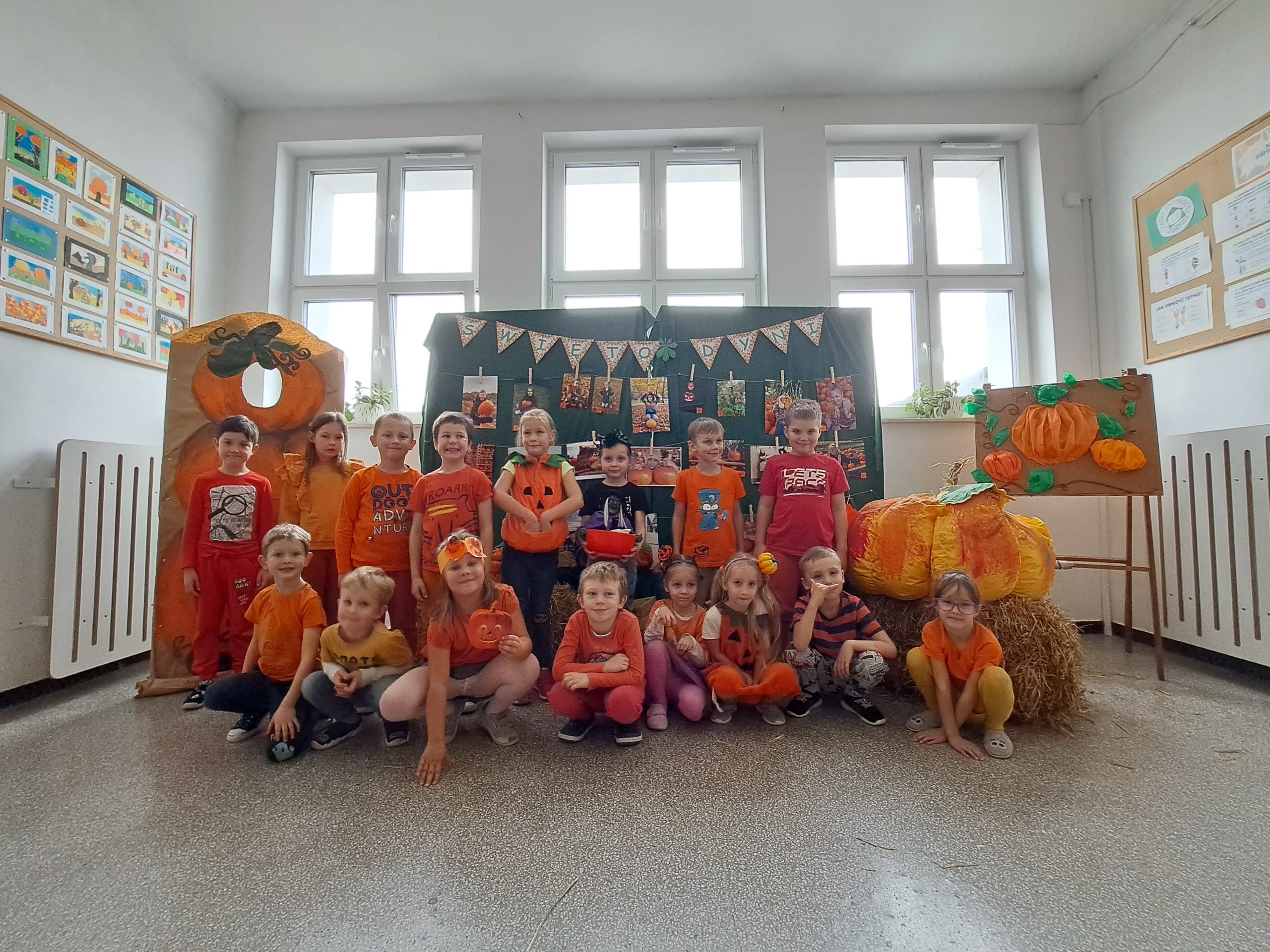 Dzieci stoją na tle granatowej tablicy, na której wiszą zdjecia przedstawiające dzieci z dyniami. Zrówkowicze są ubrani w pomarańczowe stroje na znak obchodów Dnia Dyni.