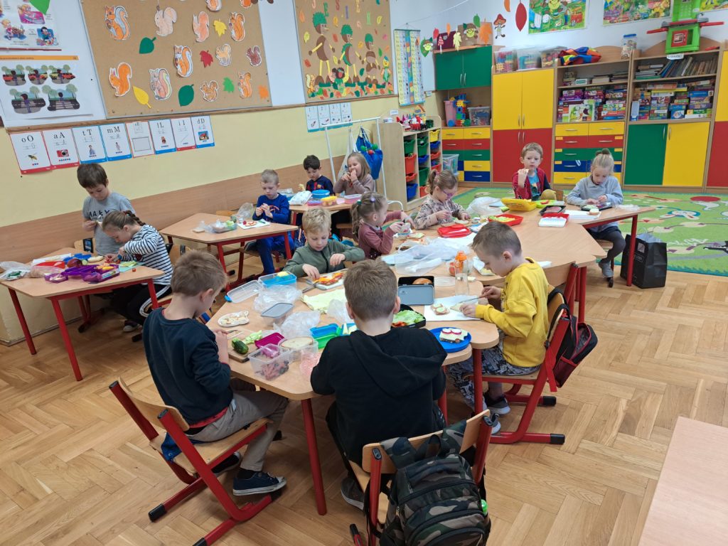  Dzieci siedzą przy stolikach, na których leżą kolorowe warzywa i inne zdrowe produkty potrzebne do przygotowanie zdrowych kanapek.