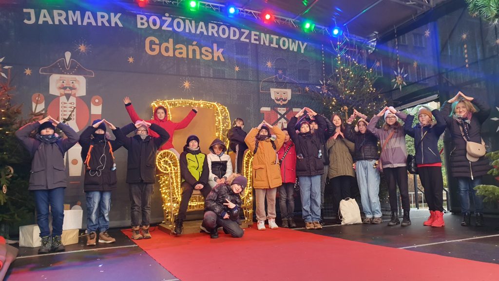 Szesnastu uczniów wraz z wychowawcą, ustawieni rzędem, trzymają ręce nad głowami na kształt choinek. dwóch chłopców siedzi na tronie. Nad dziećmi napis: Jarmark Bożonarodzeniowy Gdańsk