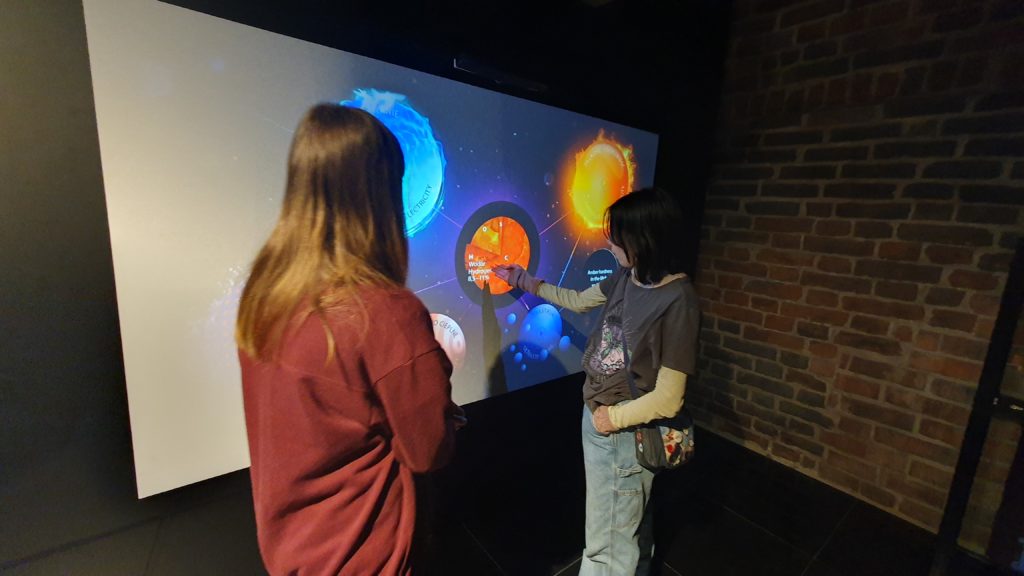 Dwie dziewczynki przed ekranem pokazującym planety. Jedna z nich wskazuje ręką środkową planetę.