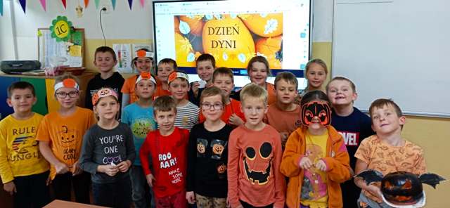 Grupa chłopców i dziewczynek ubrana na pomarańczowo, niektórzy mają na głowie opaski z papierową dynią stoją w klasie przed tablicą interaktywną na której jest duże zdjęcie dyni z napisem Dzień dyni. 
