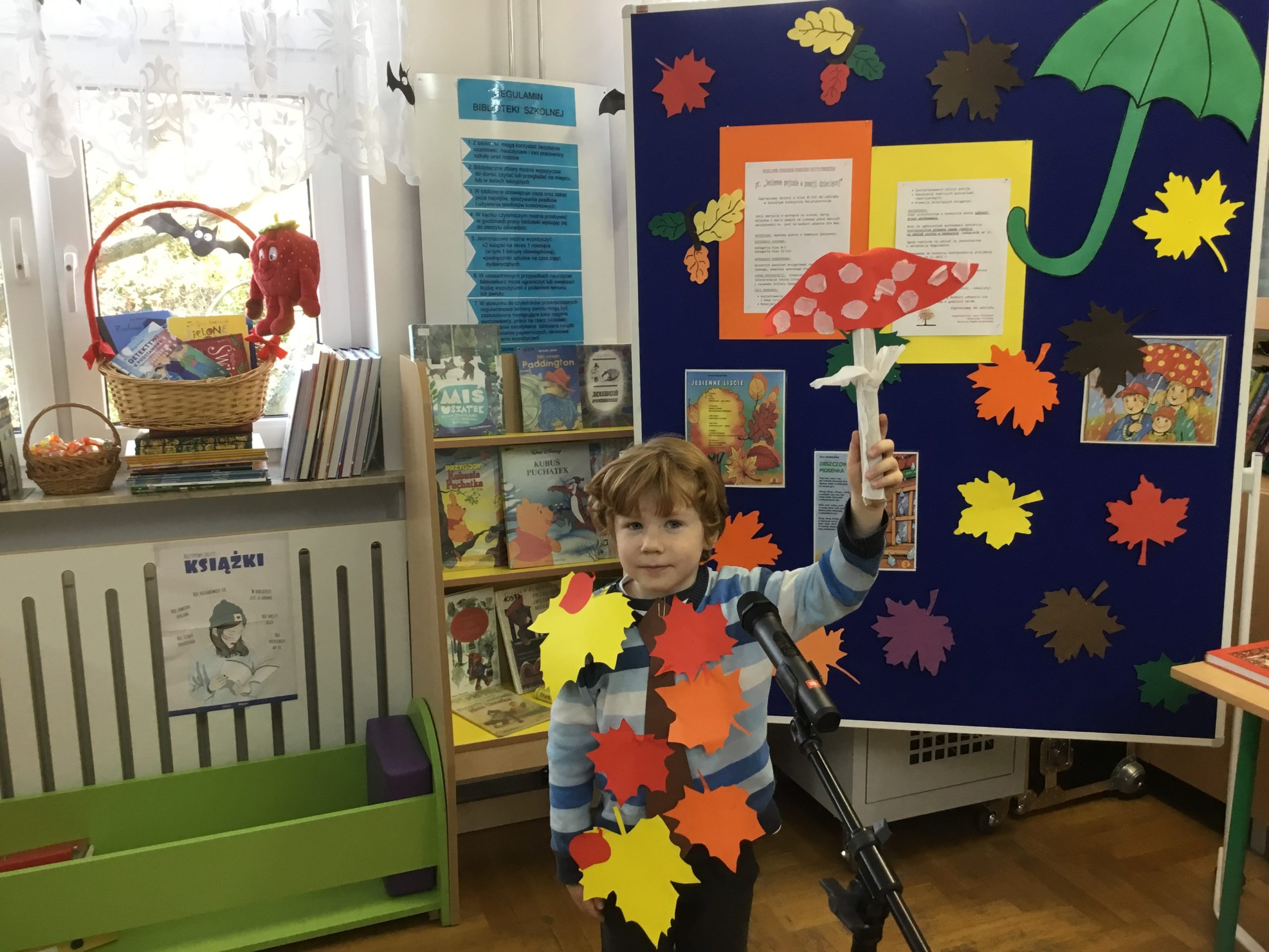 chłopiec, który orzymał 1 miejsce podczas występu w bibliotece szkolnej, w tle tablica informująca o konkursie, na parapecie kosz z nagrodami