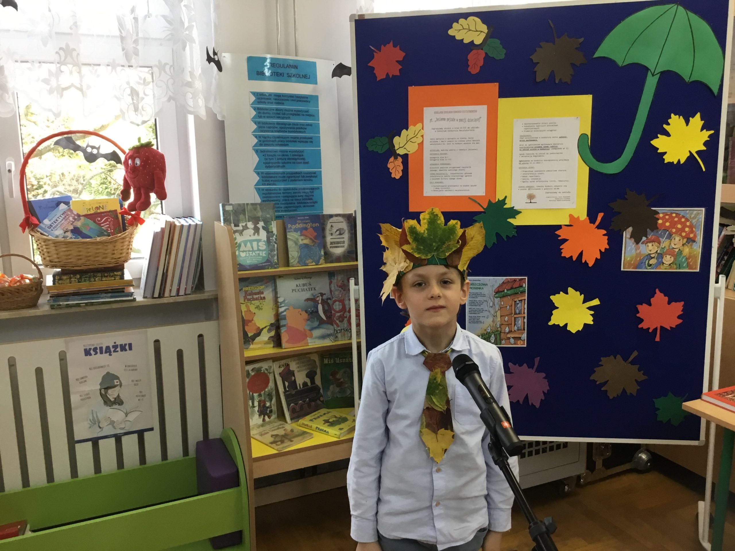 chłopiec, który orzymał 2 miejsce podczas występu w bibliotece szkolnej, w tle tablica informująca o konkursie, na parapecie kosz z nagrodami