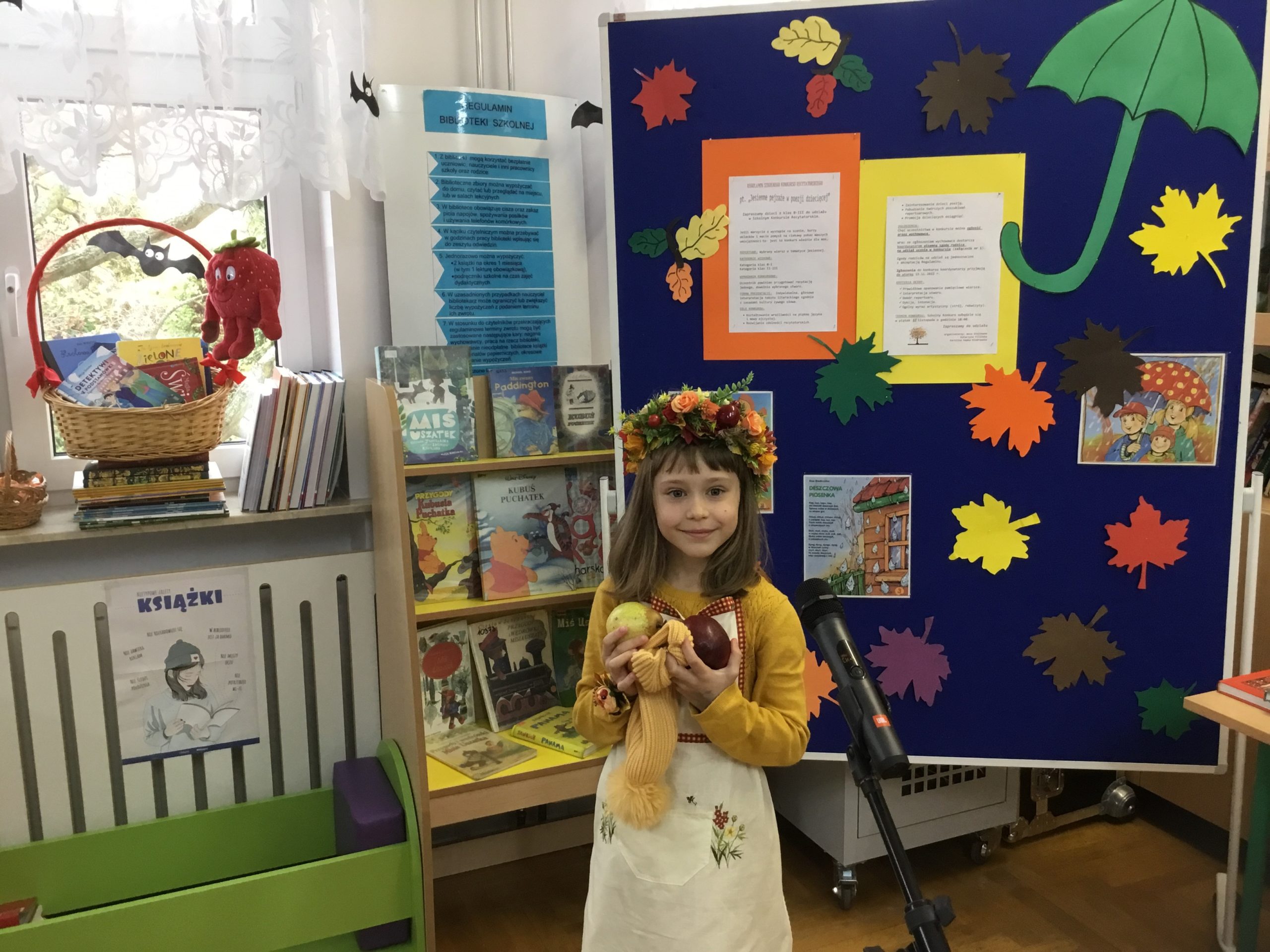 dziewczynka, która orzymała 3 miejsce podczas występu w bibliotece szkolnej, w tle tablica informująca o konkursie, na parapecie kosz z nagrodami