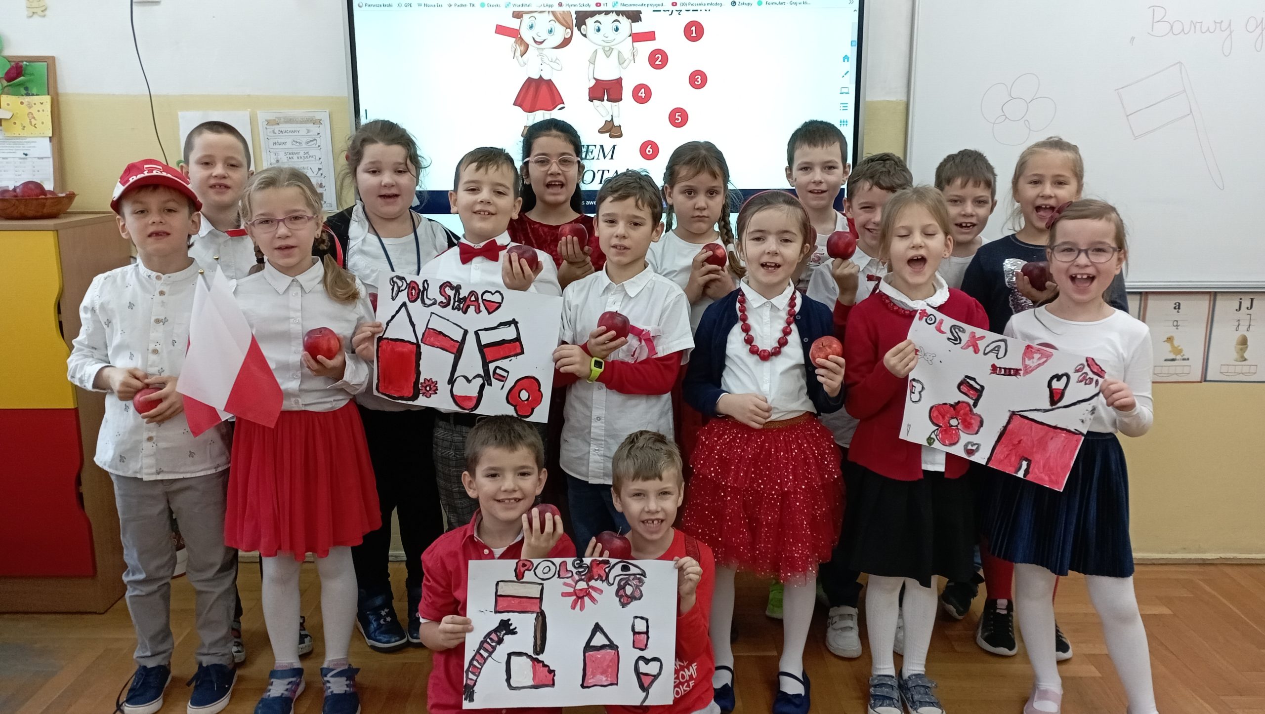 17 uczniów klasy 1c ubranych w biało- czerwone barwy stoi lub klęczy ustawionych w trzy rzędy w sali lekcyjnej na tle tablicy multimedialnej pozując do zdjęcia. W dłoniach trzymają jabłka i malunki w ojczystych barwach.