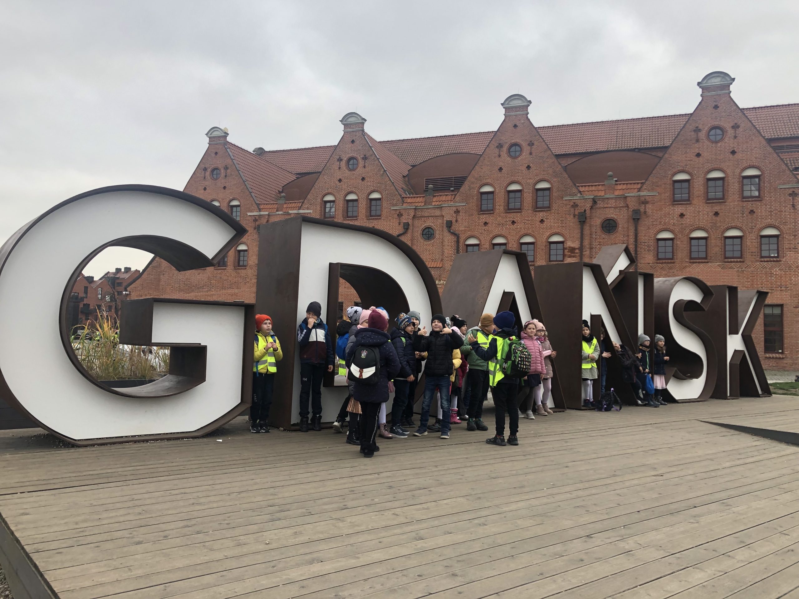 23 dzieci stoi na tle przestrzennego napisu Gdańsk. Na drugim planie budynek Filharmonii Bałtyckiej